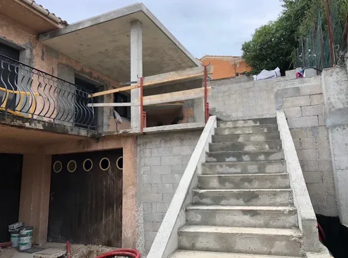 Escalier extérieur en béton menant à une maison en construction avec balcon et murs inachevés.