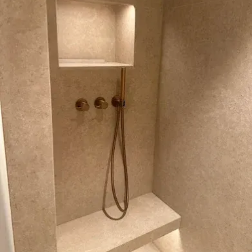 Cabine de douche carrelée beige avec étagère encastrée, deux boutons au mur et douchette à main.
