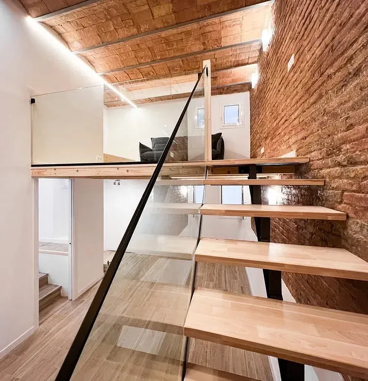 Una escalera de madera con barandilla de vidrio y una pared de ladrillo.
