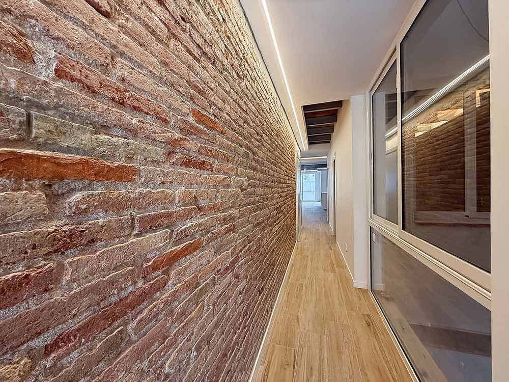 Un pasillo estrecho con una pared rústica de ladrillo rojo en un lado, suelo de madera clara y una gran ventana de cristal en el otro.