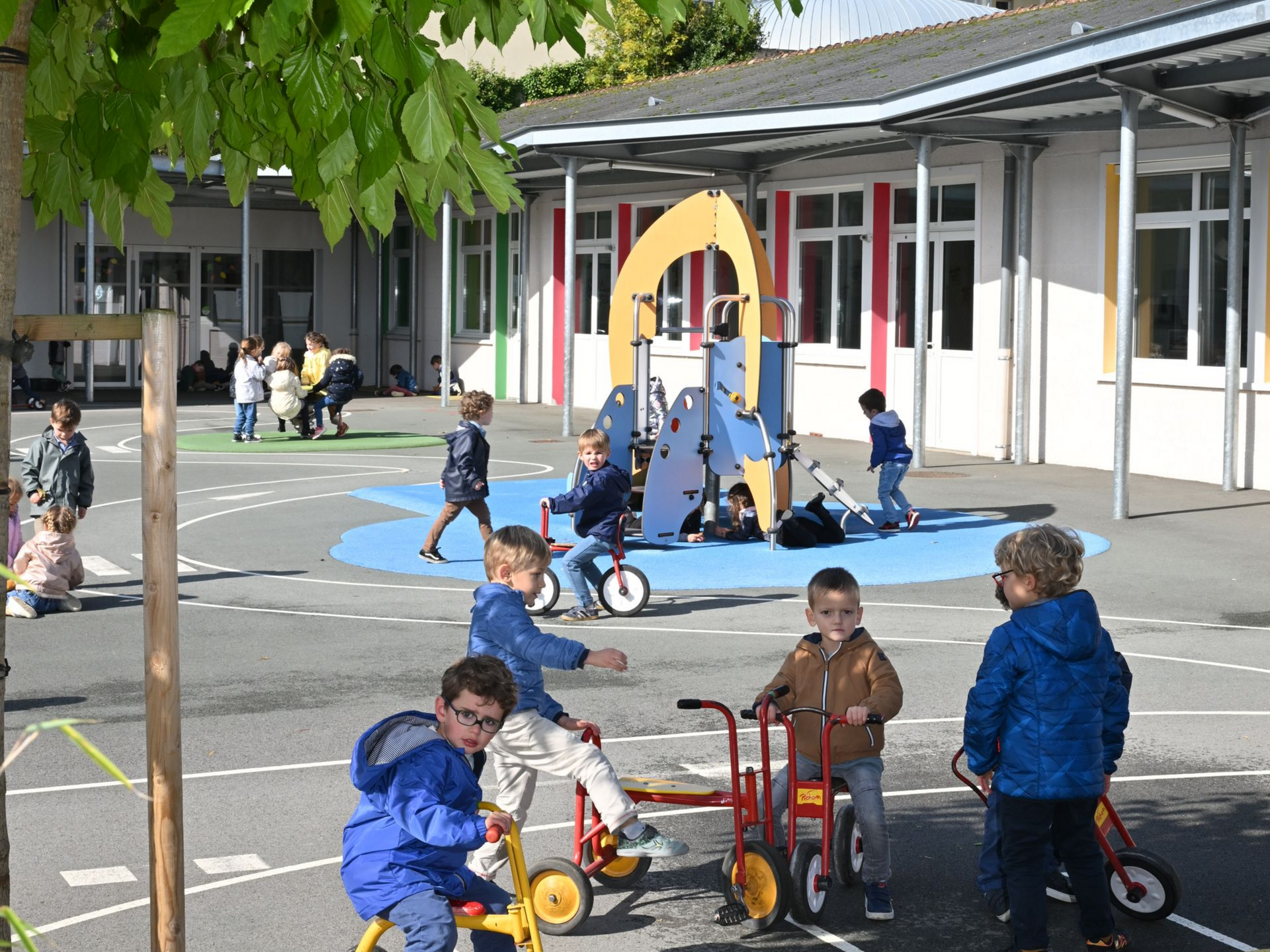 Cour d’école avec plusieurs enfants jouant, certains sur des tricycles, près d’une structure de jeu colorée au centre