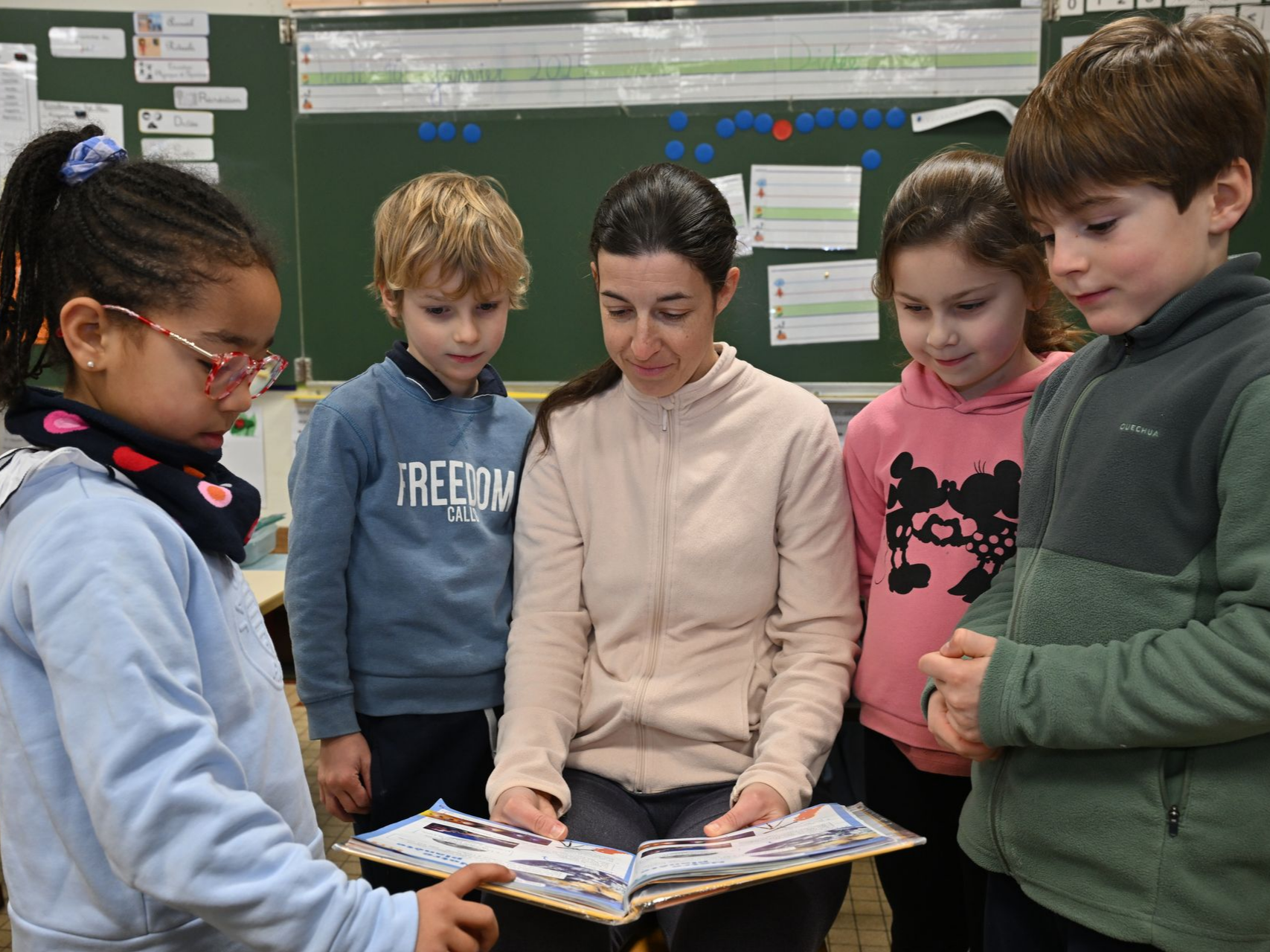 Personne assise en classe tenant un livre ouvert, entourée d’enfants debout écoutant la lecture