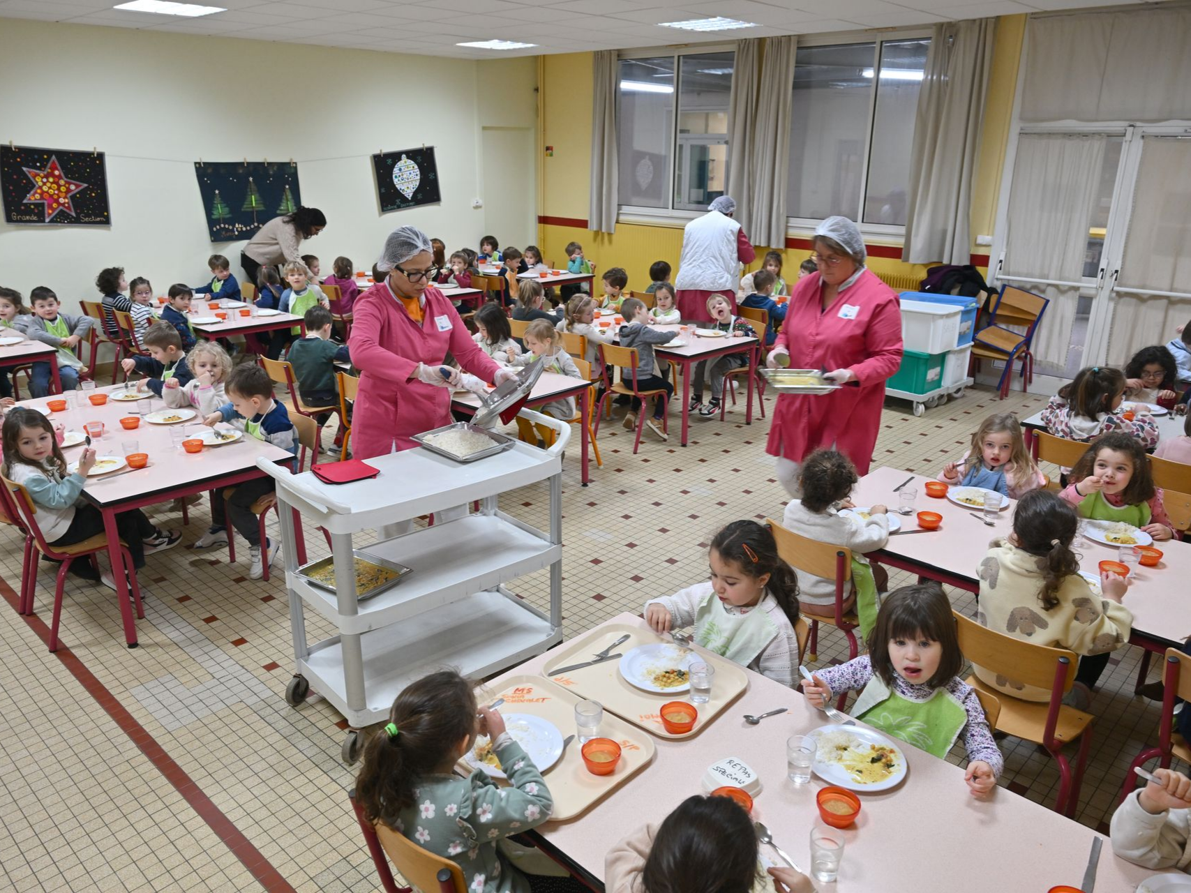 Cantine scolaire avec plusieurs tables où des enfants prennent leur repas, et du personnel en tablier rose servant les plats