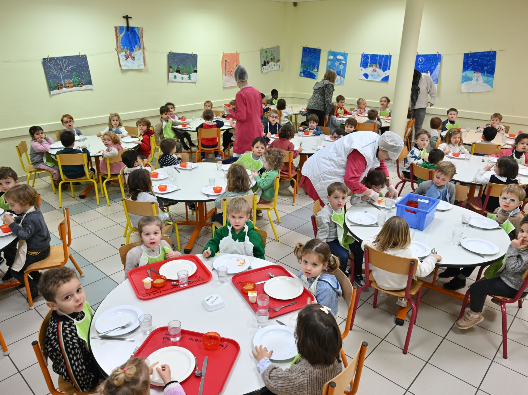 Grande salle de cantine avec plusieurs tables rondes où des enfants sont assis pour le repas