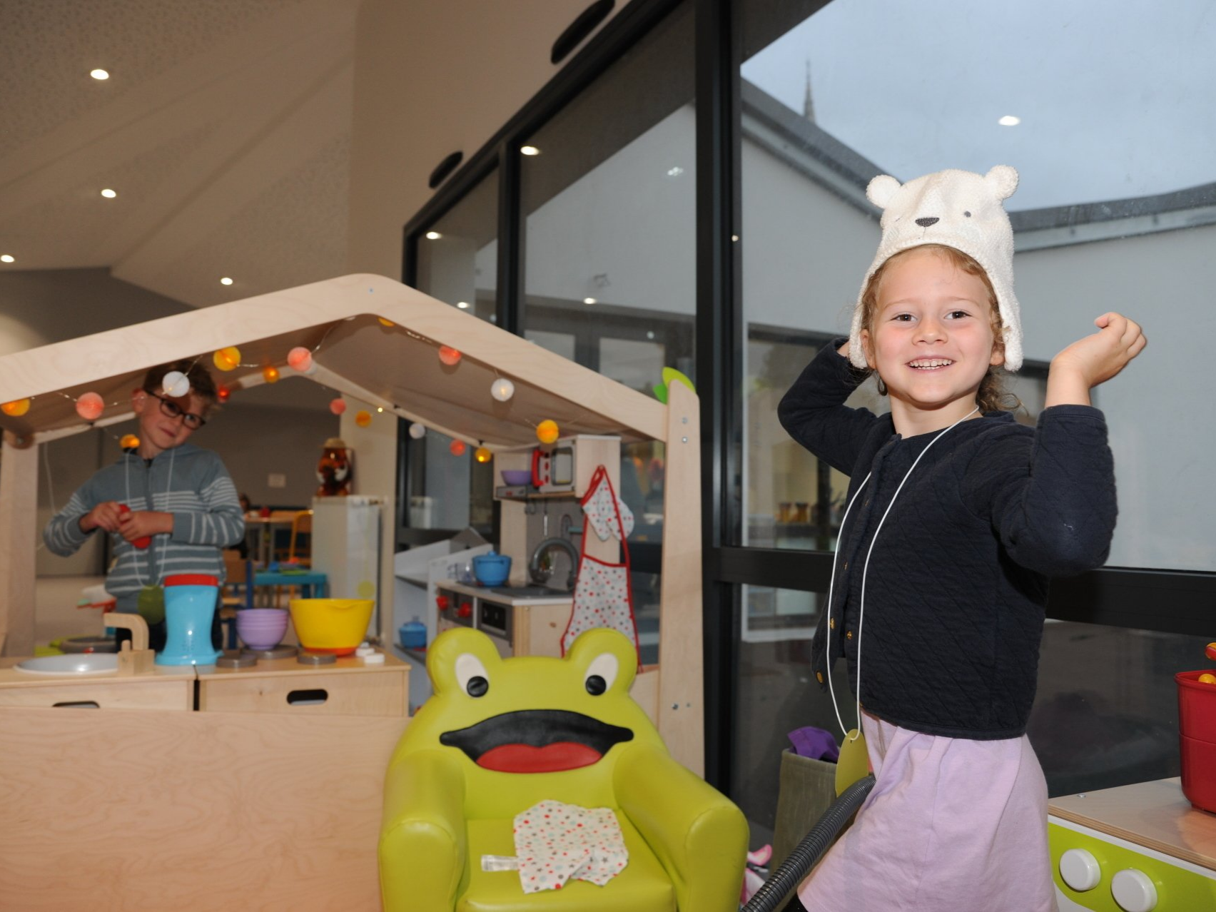 Une enfant avec un bonnet en forme d'animal près d'une maison en bois où joue un jeune garçon à lunettes