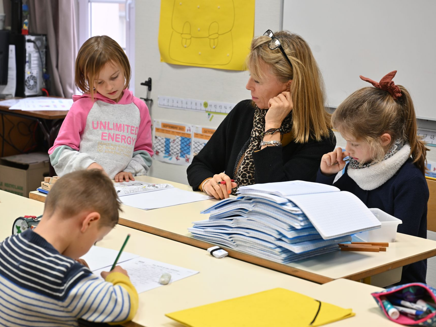 Enseignante assise à une table avec plusieurs cahiers ouverts, entourée d’enfants travaillant sur des exercices