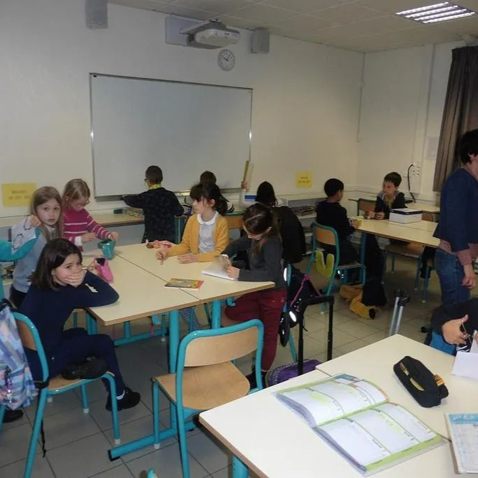 Des enfants assis à leurs pupitres dans une salle de classe, écrivant ou regardant des feuilles ; un tableau blanc en arrière-plan.