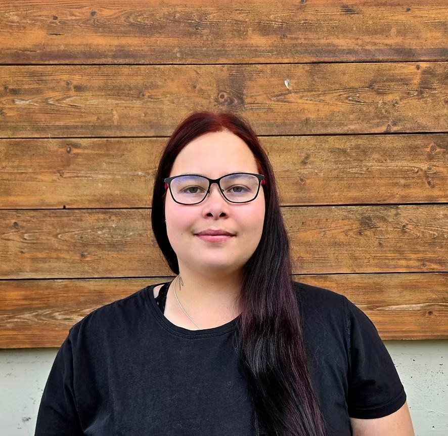 Frau mit Brille und dunklem Haar, die ein schwarzes Hemd trägt, vor einem Holzhintergrund.