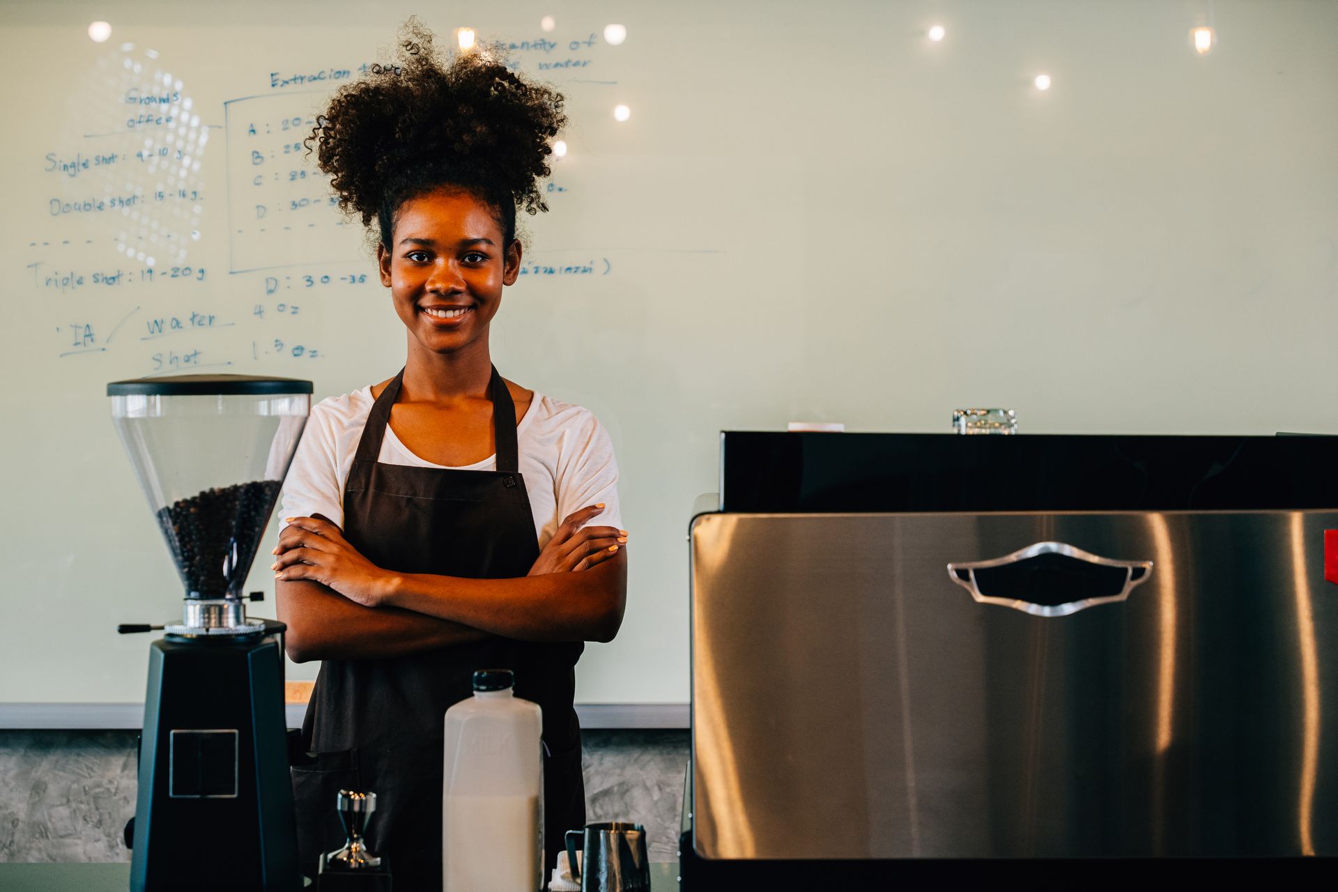 Une barista, debout derrière sa machine à café dans un café moderne, sourit, les bras croisés.