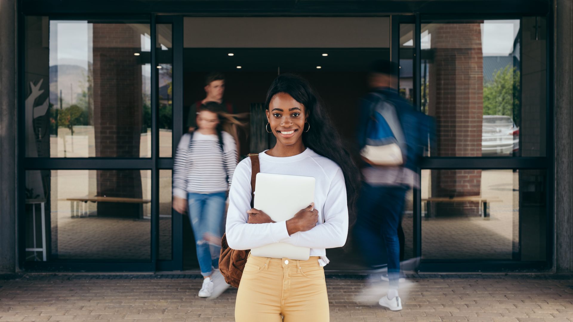 Une étudiante, les bras croisés, se tient devant l'entrée de son école, ses camarades de classe flous derrière elle.