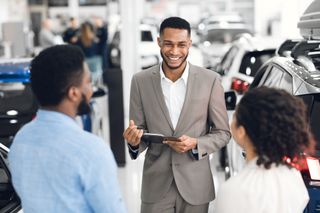 Un vendeur de voitures souriant présente une tablette à deux clients dans un showroom lumineux.