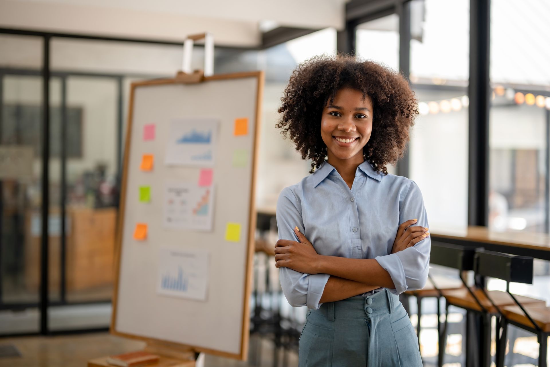 Une personne souriante, les bras croisés, dans un bureau à côté d'un tableau blanc couvert de post-it.