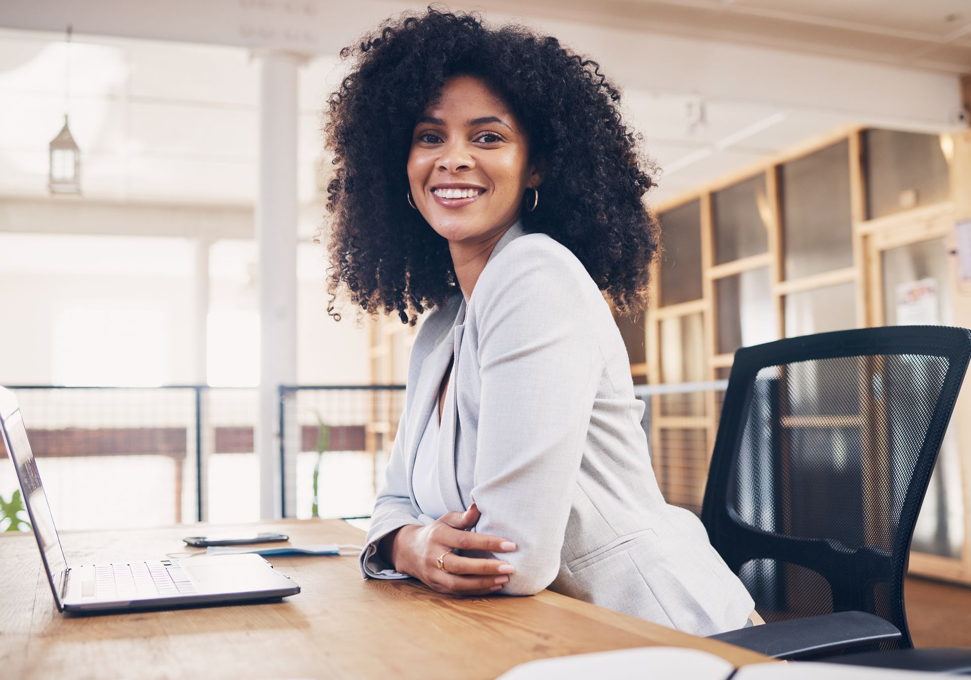 Une femme souriante est assise à un bureau dans un bureau lumineux, avec un ordinateur portable et une chaise à proximité.