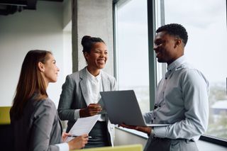 Trois collègues sourient et discutent dans un bureau lumineux, l'un tenant un ordinateur portable et l'autre des papiers.