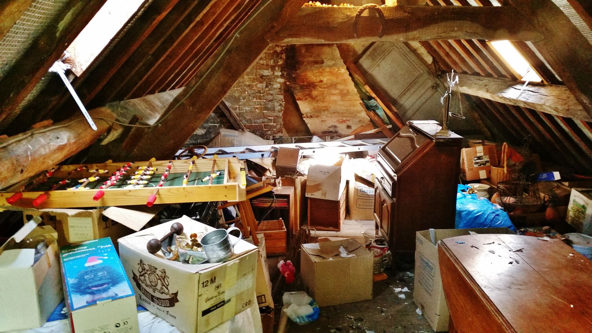 Grenier encombré de poutres en bois, de cartons, d'un baby-foot et d'un piano. La lumière naturelle y pénètre abondamment.