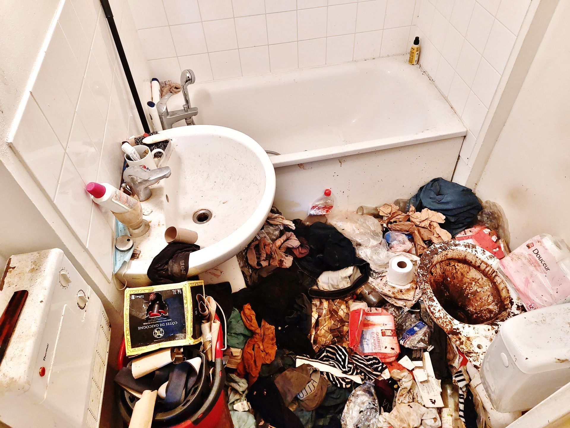 Salle de bain encombrée : lavabo, baignoire et piles de vêtements et d’objets sur le sol.