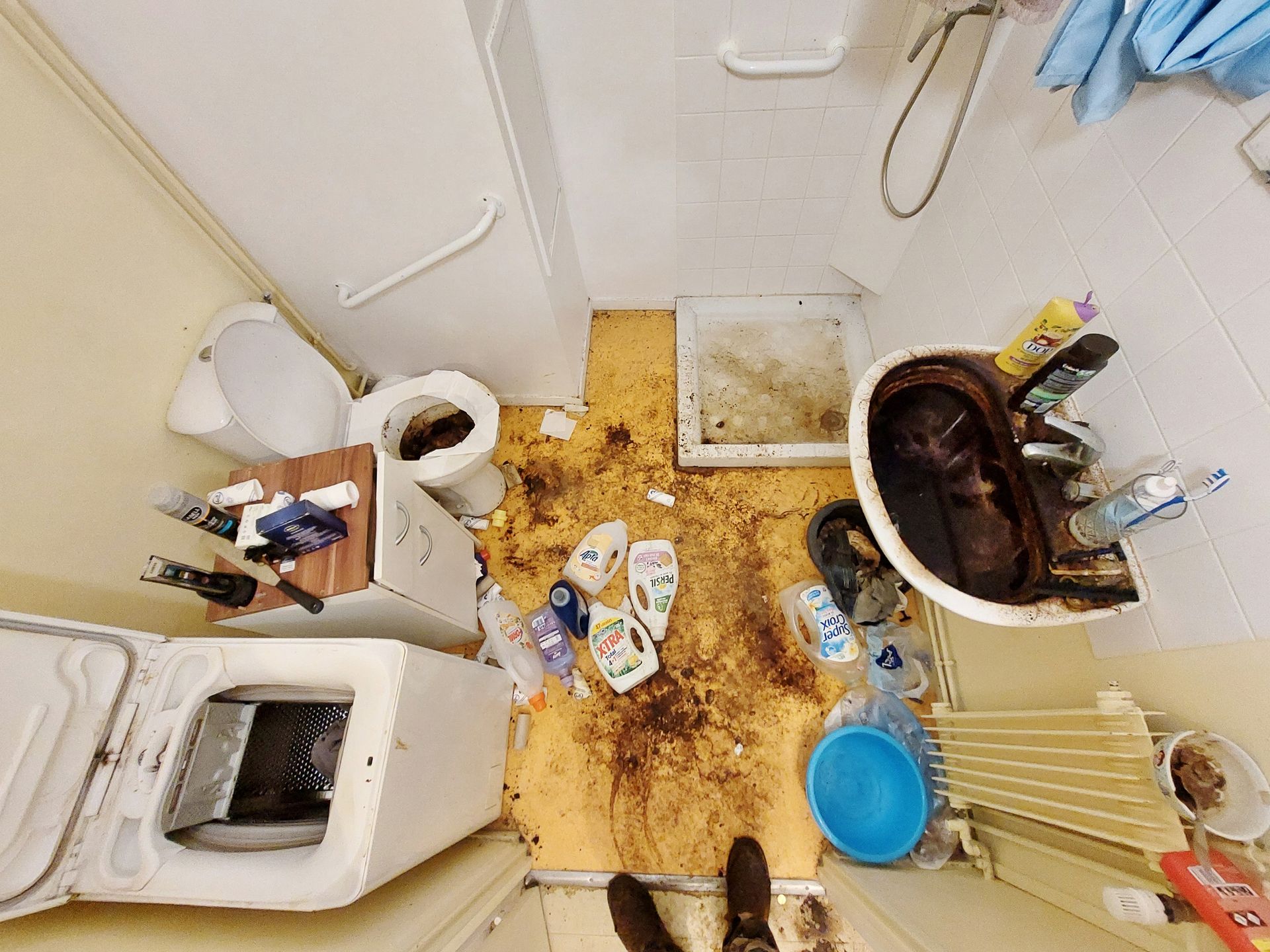 Salle de bain sale avec des ordures et des débris au sol, comprenant des toilettes, une baignoire et une machine à laver.