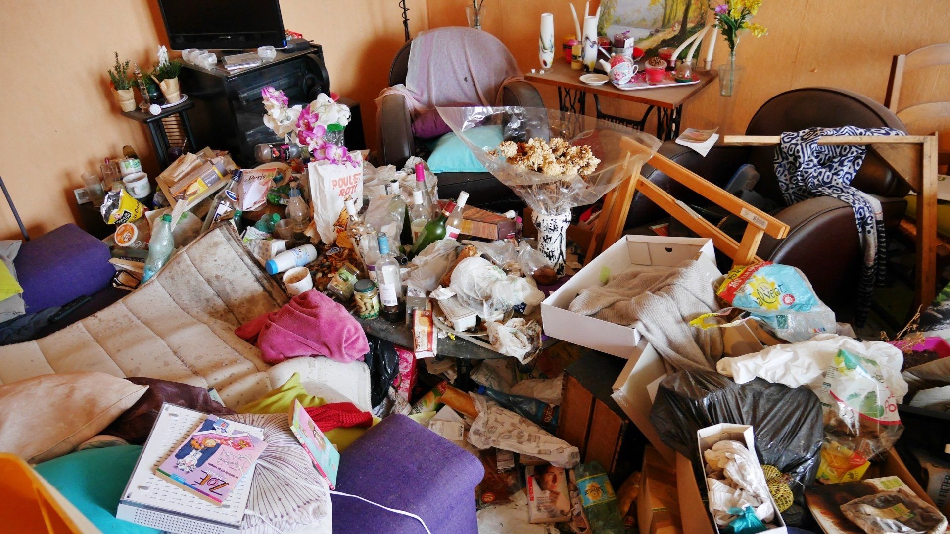 Un salon avec une table et des fauteuils recouverts d'objets et déchets.