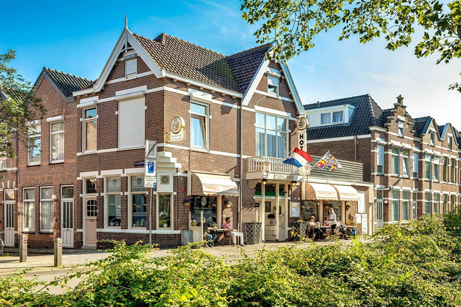 Bruin bakstenen gebouw met luifels, zitgelegenheid op de stoep en een Nederlandse vlag.