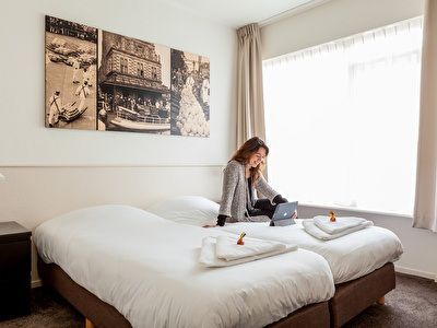 Vrouw zit op een hotelbed, met een laptop in haar hand, bij een raam, met kunst boven het hoofdeinde.