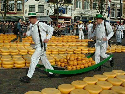 Mannen in witte gewaden en hoeden racen met een slee vol kaas door een markt, omringd door vele kaaswielen.