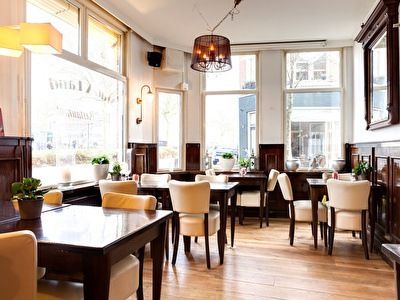 Restaurantinterieur met tafels, stoelen en grote ramen. Donkere houten lambrisering en lichtgekleurde stoelen.