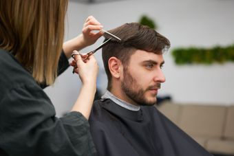 Un coiffeur coupe les cheveux d'un homme aux ciseaux. Homme assis, vêtu d'une cape de barbier. Cheveux blonds, ambiance neutre.
