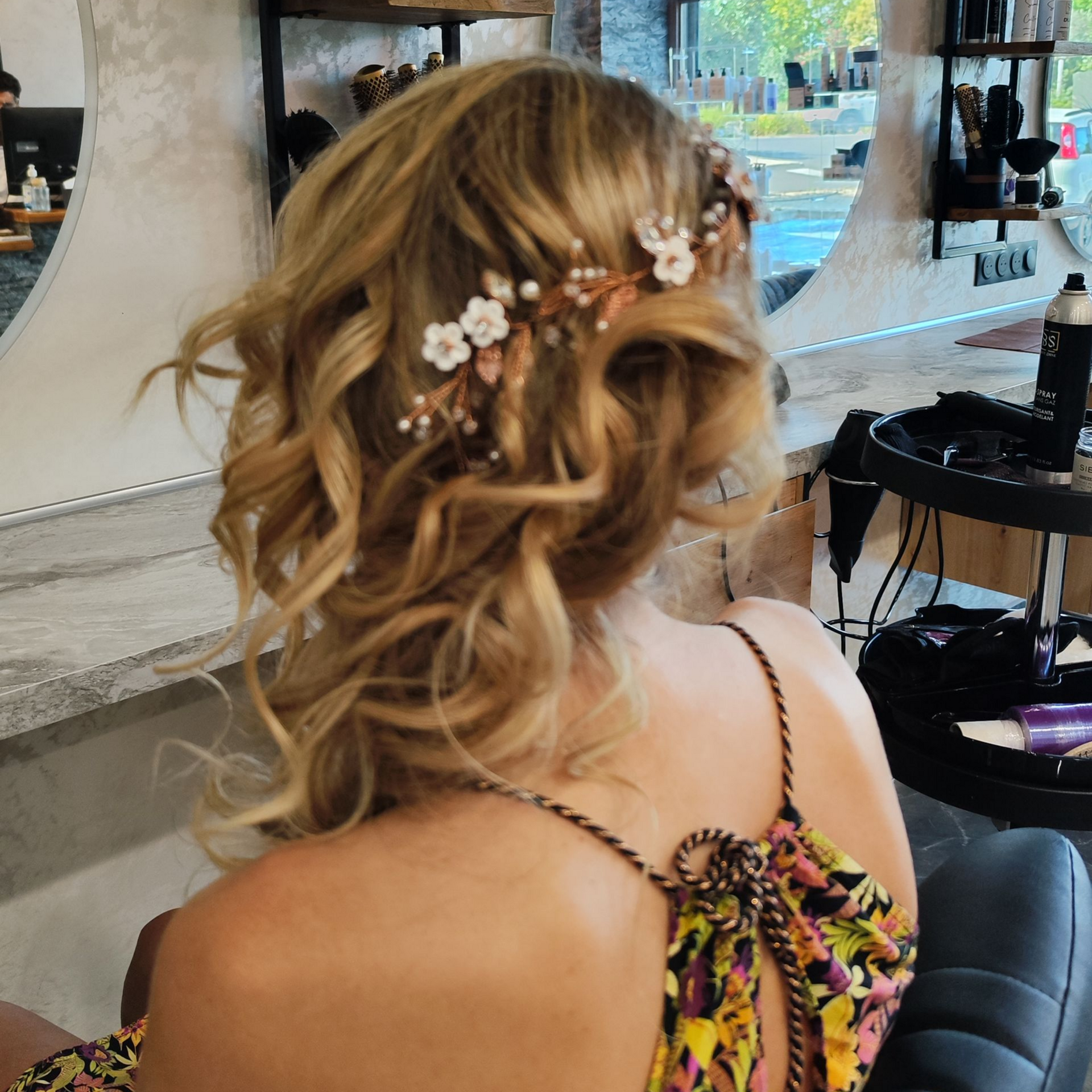 Femme aux cheveux blonds ondulés, coiffée d'une barrette à fleurs, dans un salon de coiffure.