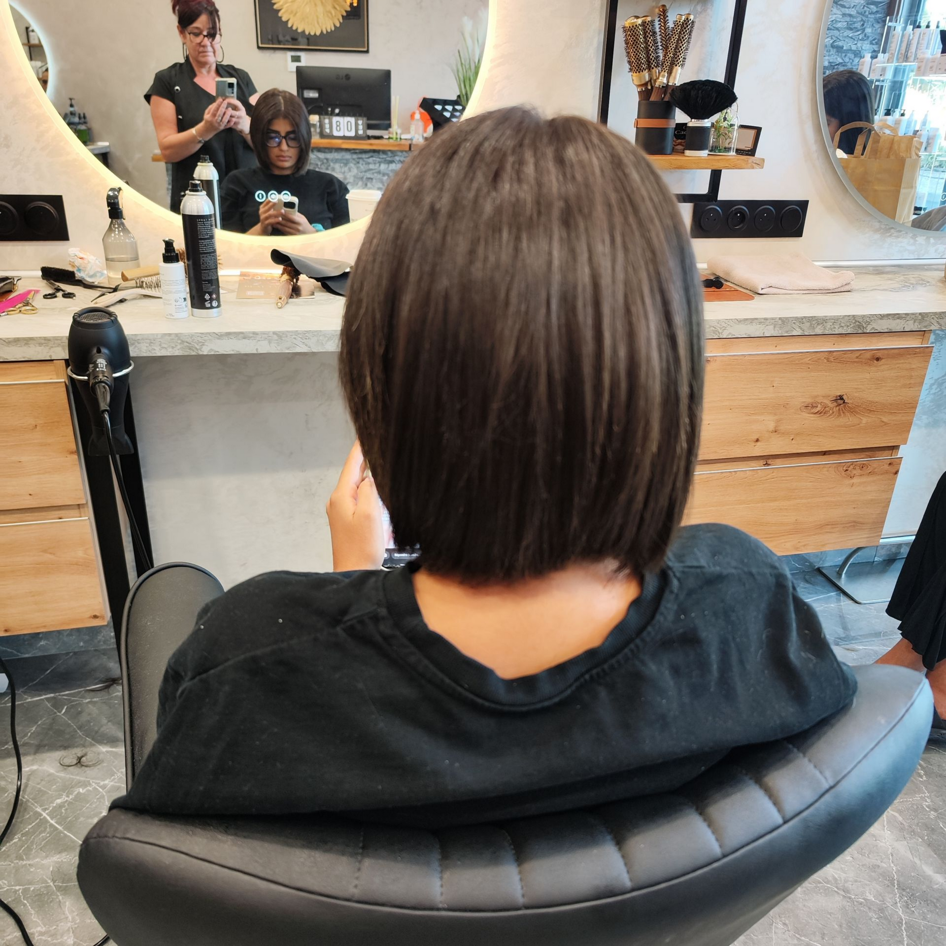 Femme assise sur un fauteuil de salon, les cheveux coupés au carré, vue de dos. On aperçoit une coiffeuse et un miroir.