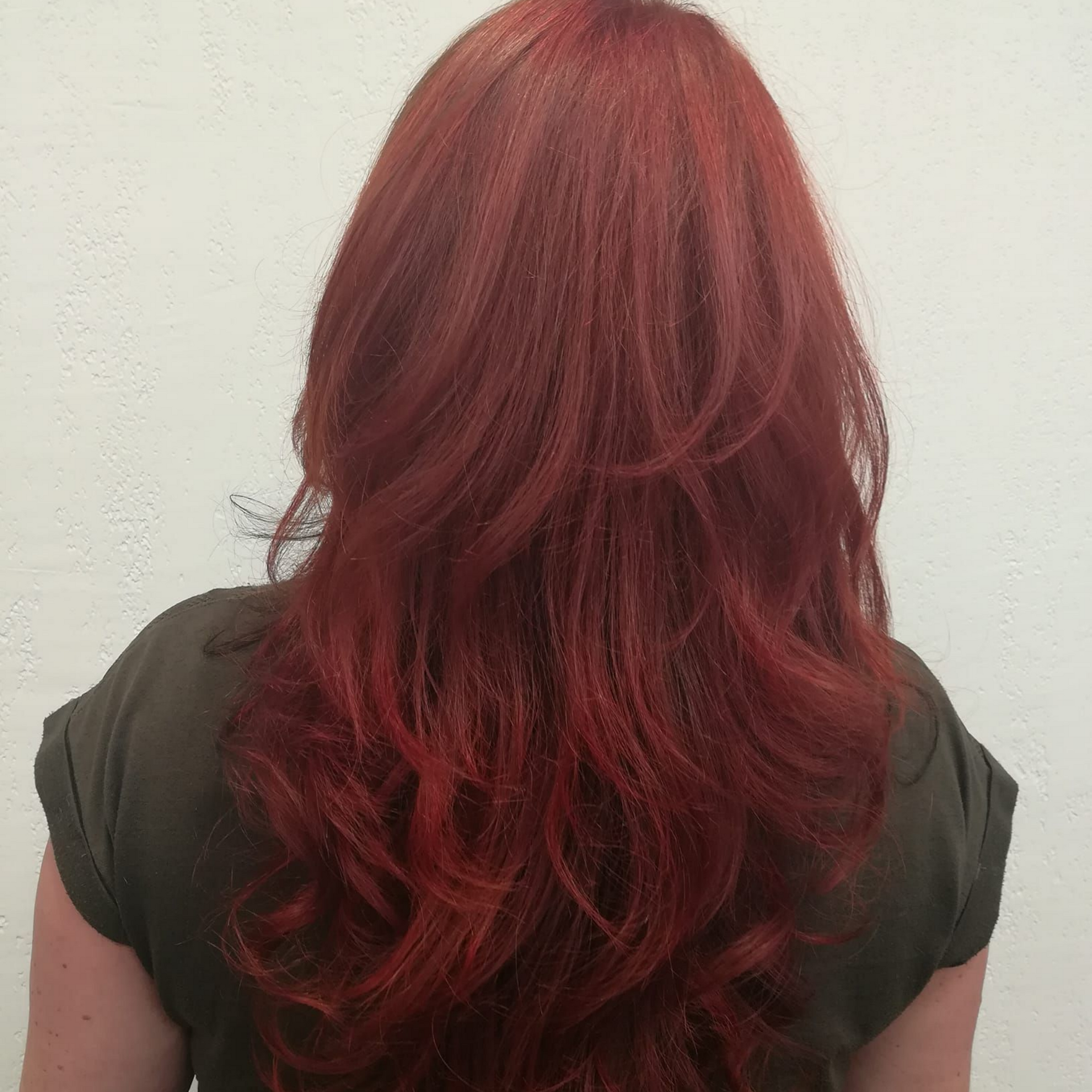 Femme aux longs cheveux roux dégradés, portant une chemise verte, devant un mur blanc.