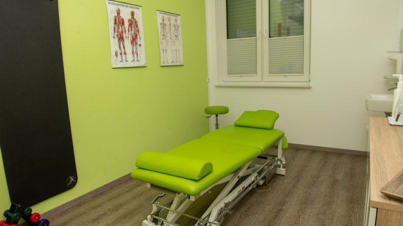 In einem Raum neben einem Fenster steht ein grüner Massagetisch.