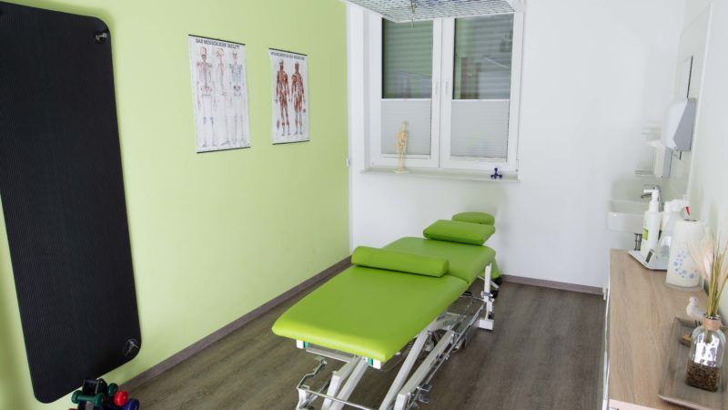 In einem Raum neben einem Fenster steht ein grüner Massagetisch.