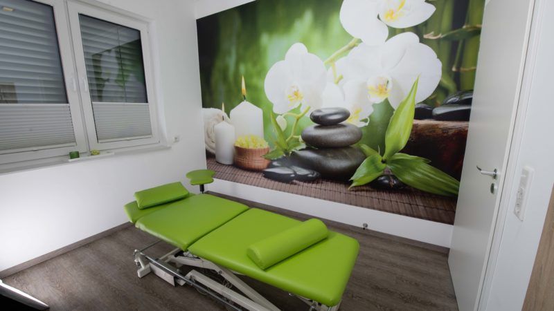 Ein grüner Massagetisch in einem Raum mit einem Bild von Blumen und Steinen an der Wand.