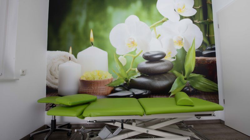 Ein grüner Massagetisch in einem Raum mit einem Bild von Blumen und Steinen an der Wand.