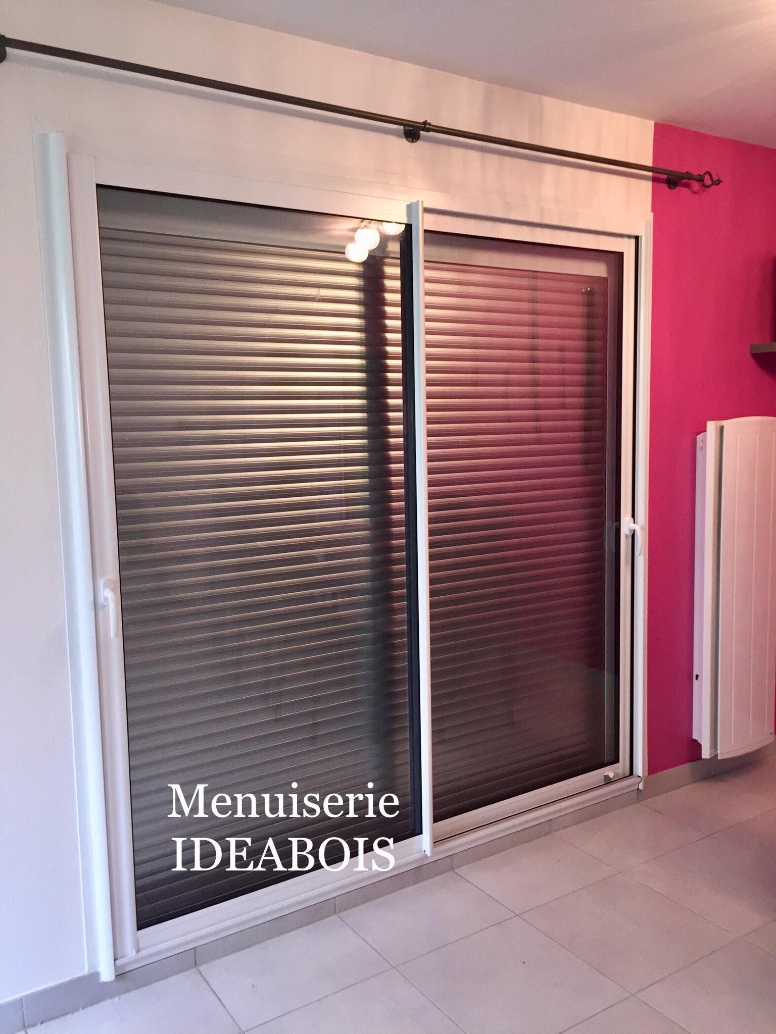 Porte coulissante en verre à cadre blanc avec stores gris fermés et tringle à rideaux noire ; mur rose à droite.