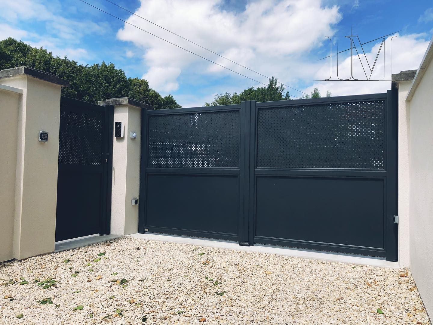 Portail en métal gris foncé, encadré de murs beige clair. Sol gravillonné, ciel bleu en arrière-plan.