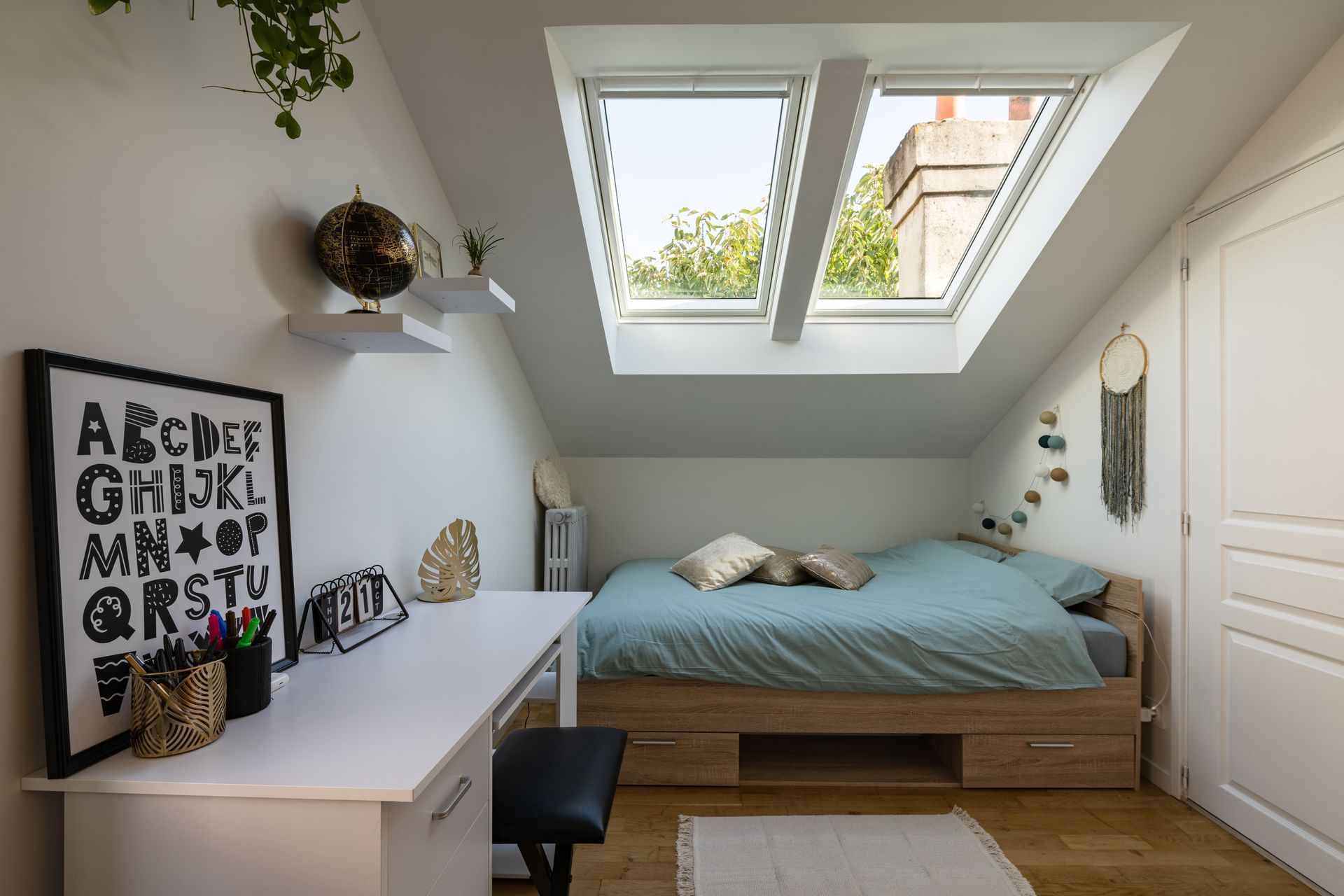 Chambre mansardée confortable avec bureau, lit et puits de lumière. Murs blancs, parquet et éléments décoratifs.