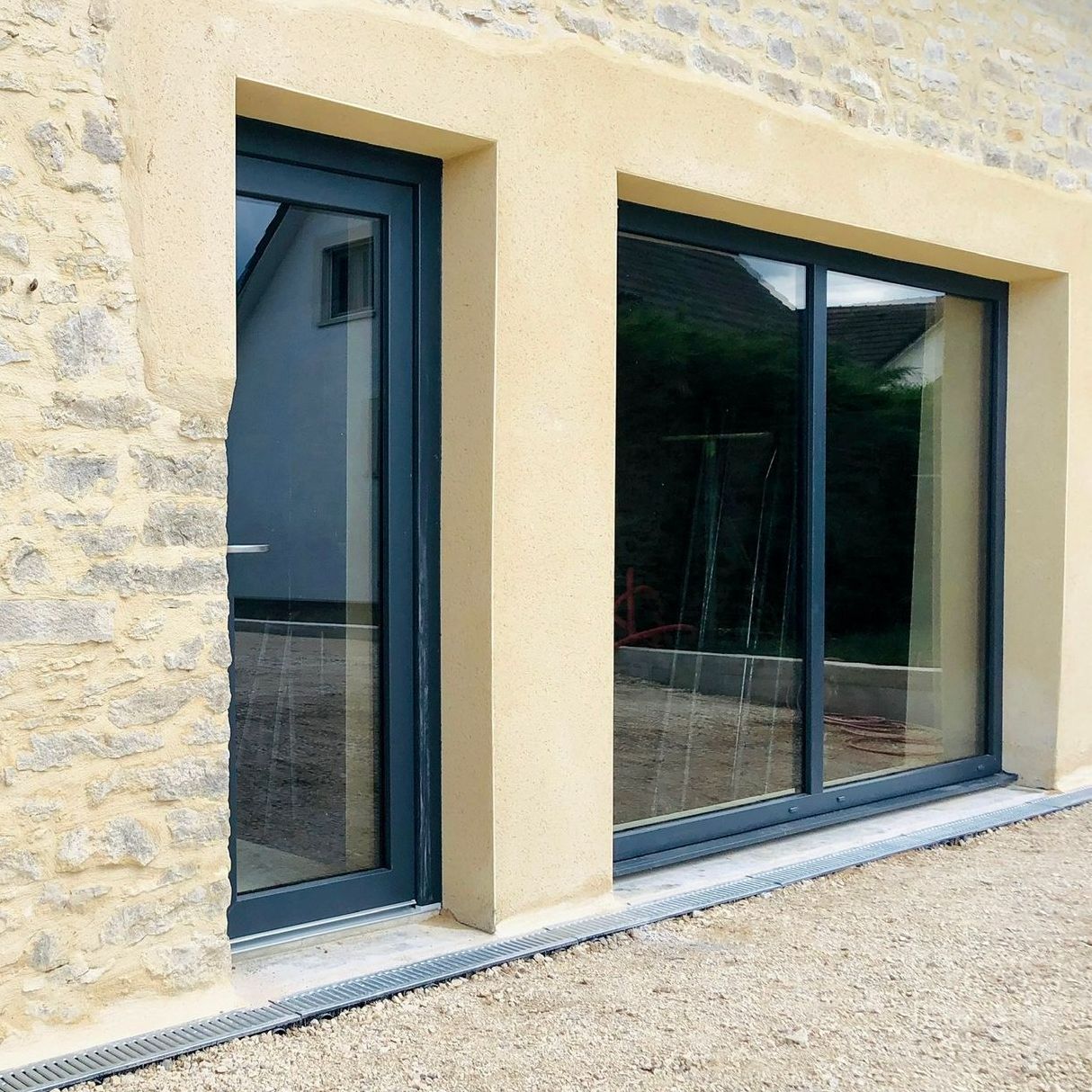 Porte à cadre sombre et fenêtre coulissante dans un mur en stuc beige, à côté d'un mur en pierre, sur du gravier.