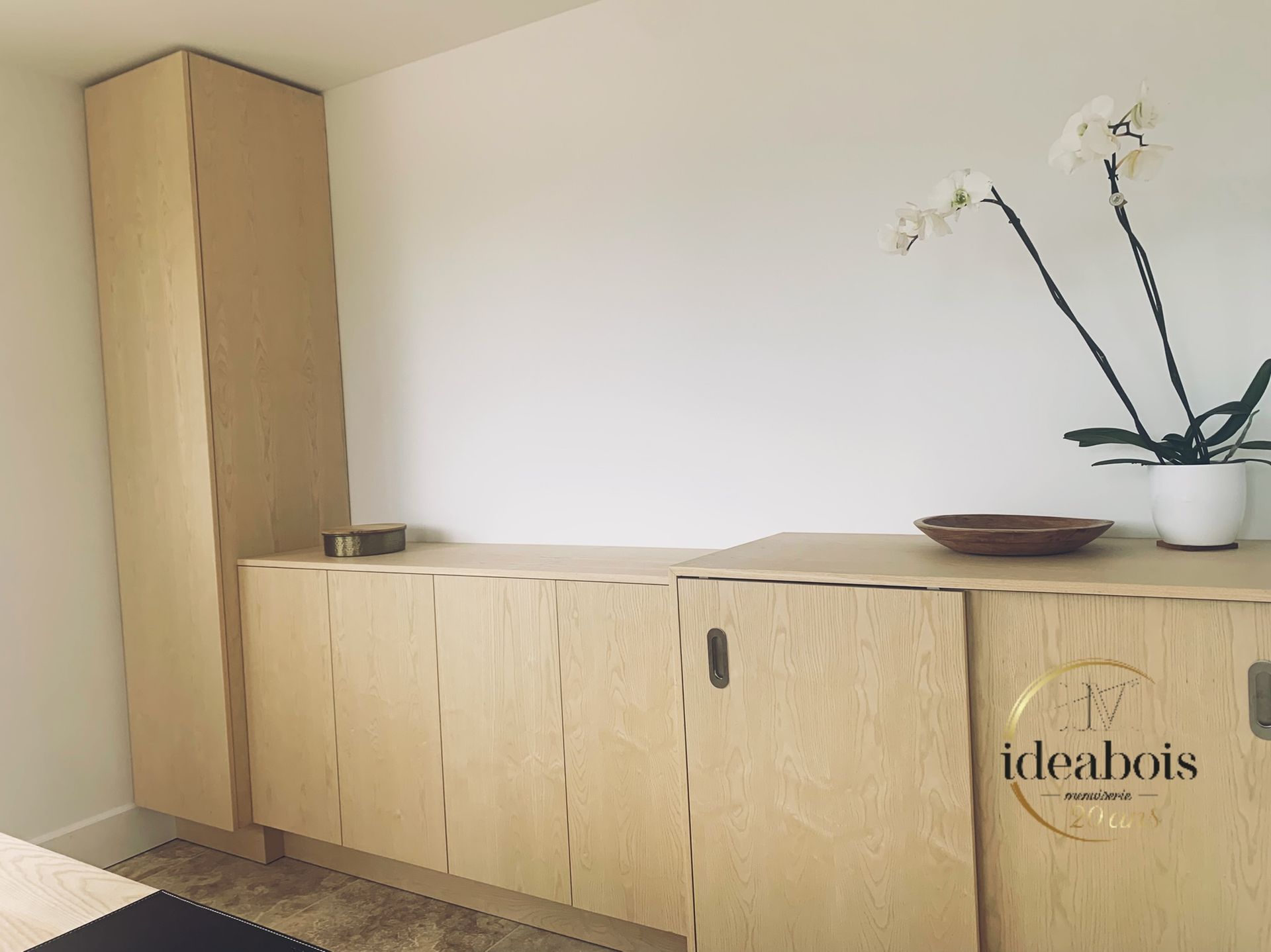 Armoire de rangement en bois clair avec armoire haute intégrée contre un mur blanc ; orchidée, bol sur le dessus.