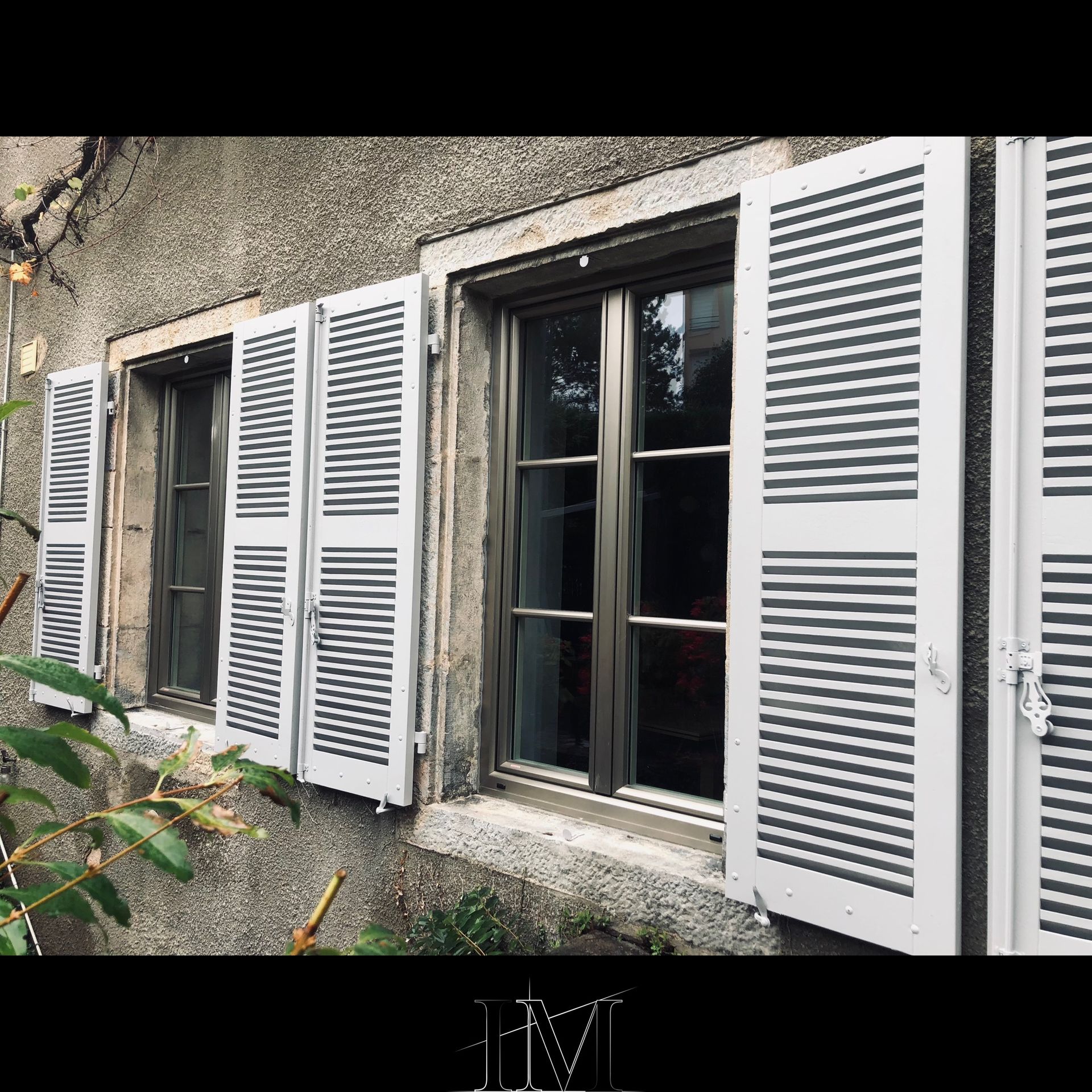 Trois fenêtres à persiennes blanches sur un mur de pierre grise, avec de la verdure au premier plan.