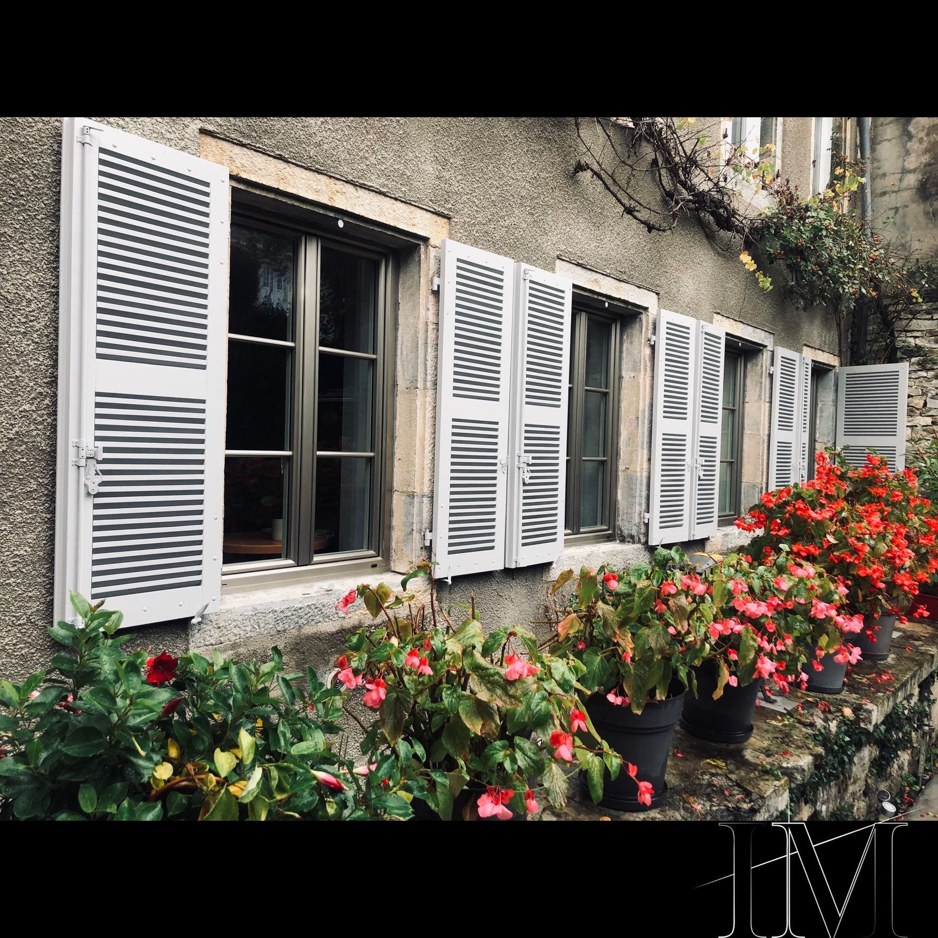 Rangée de fenêtres à volets blancs, pots de fleurs rouges et plantes grimpantes sur un bâtiment en pierre.