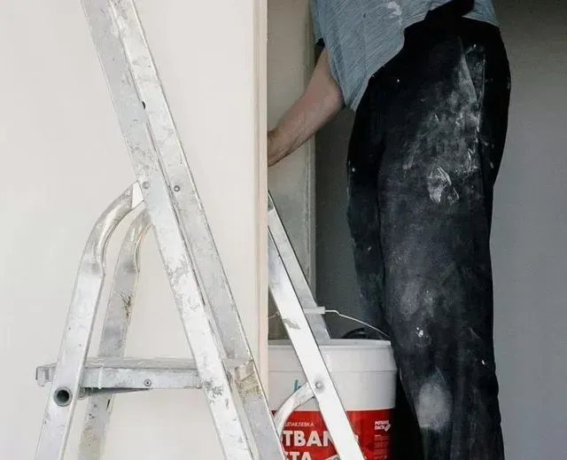 Persona pintando una pared, subida a una escalera. Cubo de pintura cerca, con pantalones manchados de pintura.