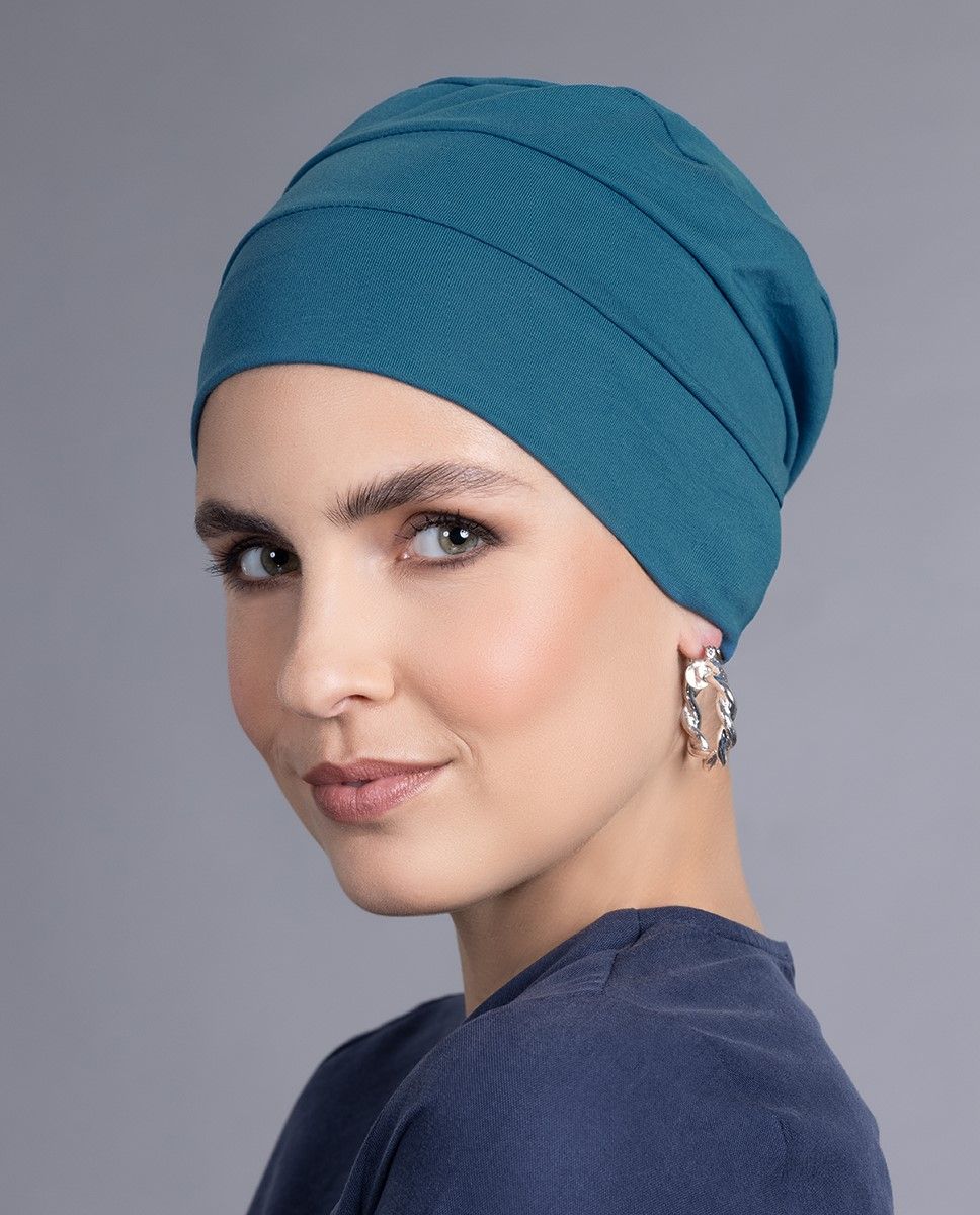 Frau mit blaugrünem Turban, lächelnd vor grauem Hintergrund. Sie trägt ein dunkelblaues Hemd und Creolen.