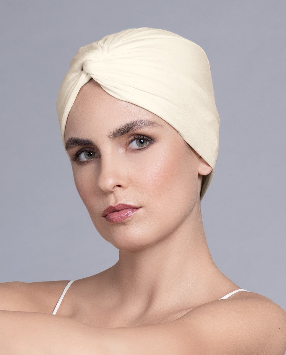 Frau mit cremefarbenem Turban, die mit neutralem Gesichtsausdruck nach vorne blickt.