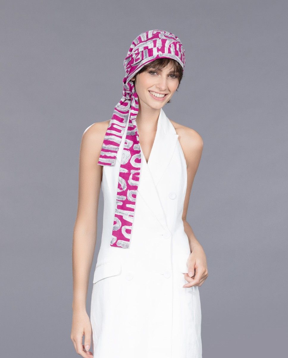 Frau in einem weißen Kleid mit rosa gemusterter Kopfbedeckung, lächelnd. Grauer Hintergrund.