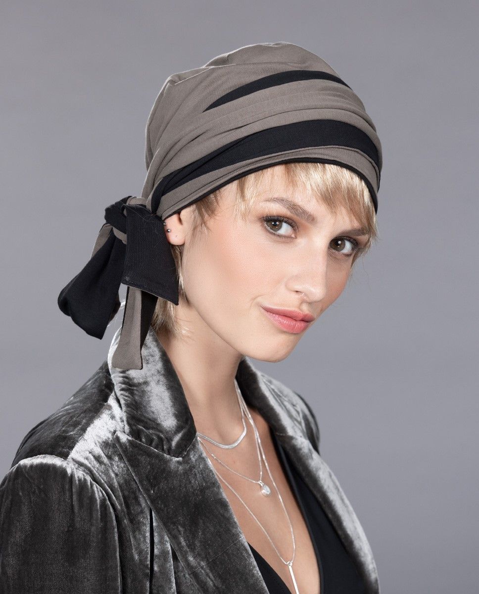 Frau trägt ein graues Kopftuch mit schwarzen Akzenten, eine schwarze Jacke und mehrere Halsketten und lächelt.