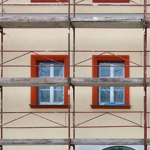 Andamio en un edificio con dos ventanas, marcos rojos, recubiertos de plástico, paredes de color beige.