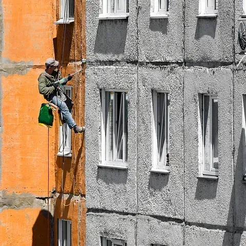 Hombre haciendo rapel en edificio, fachada naranja y gris, pintando exterior.