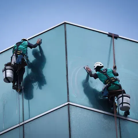 Dos limpiadores de ventanas con cuerdas limpian la esquina de vidrio de un edificio alto.