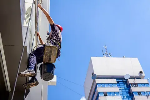 Hombre con cuerdas lavando un edificio. Cielo azul, casco, cubo y el edificio vecino al fondo.